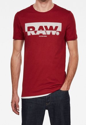 T-shirt en coton rouge avec un audacieux logo blanc "RAW." sur la poitrine. Manches courtes et col rond standard. Coupe décontractée.