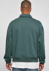 Tamnozelena oversized dukserica s kragnom, rebrastim manžetama i porubom. Mekan materijal, minimalni šavovi i jednostavan dizajn.