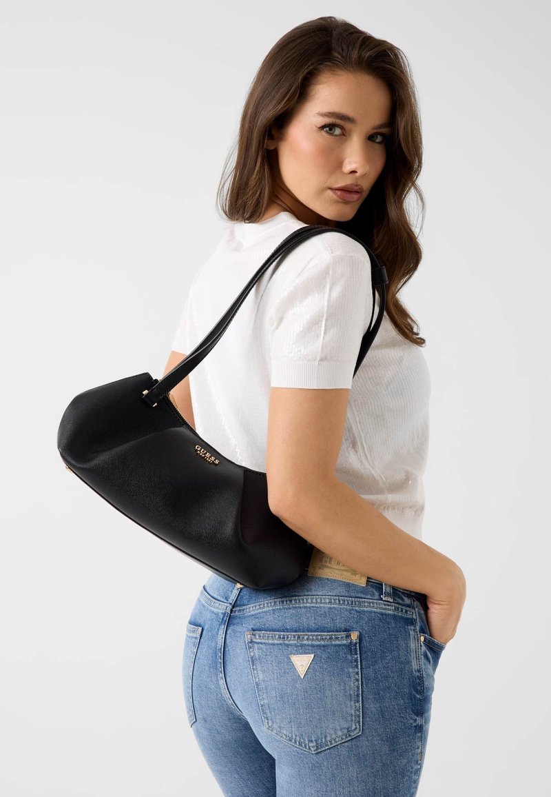 Femme portant un haut blanc à manches courtes et un jean bleu, avec un sac à épaule Guess noir avec logo doré, posant avec la main dans la poche.