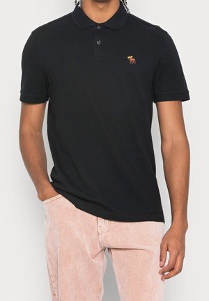 Man wearing a black short-sleeve polo shirt with a small embroidered moose logo, paired with light pink striped pants.