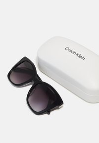 Čierne slnečné okuliare s tmavými gradientovými sklami, ostrý tvar mačacích očí, spolu s bielym tvrdým puzdrom značky "Calvin Klein." Hladký povrch.