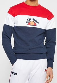 Mikina s kulatým výstřihem, blokovaná v červené, bílé a námořnické modré barvě, s ribbed manžetami a vyšitým logem na hrudi. Měkká textura.