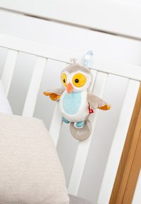 Jouet pour bébé en forme de chouette douce avec de grands yeux jaunes et un bec orange, suspendu aux barreaux blancs d'un berceau à côté d'un oreiller beige.