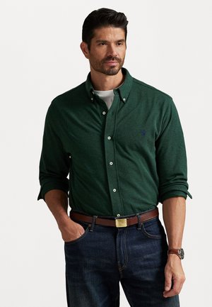 Man met kort, donker haar en baard, gekleed in een donkergroene knoopshirt over een wit T-shirt, blauwe jeans, een bruine riem en een horloge, staand met zijn hand in zijn zak.