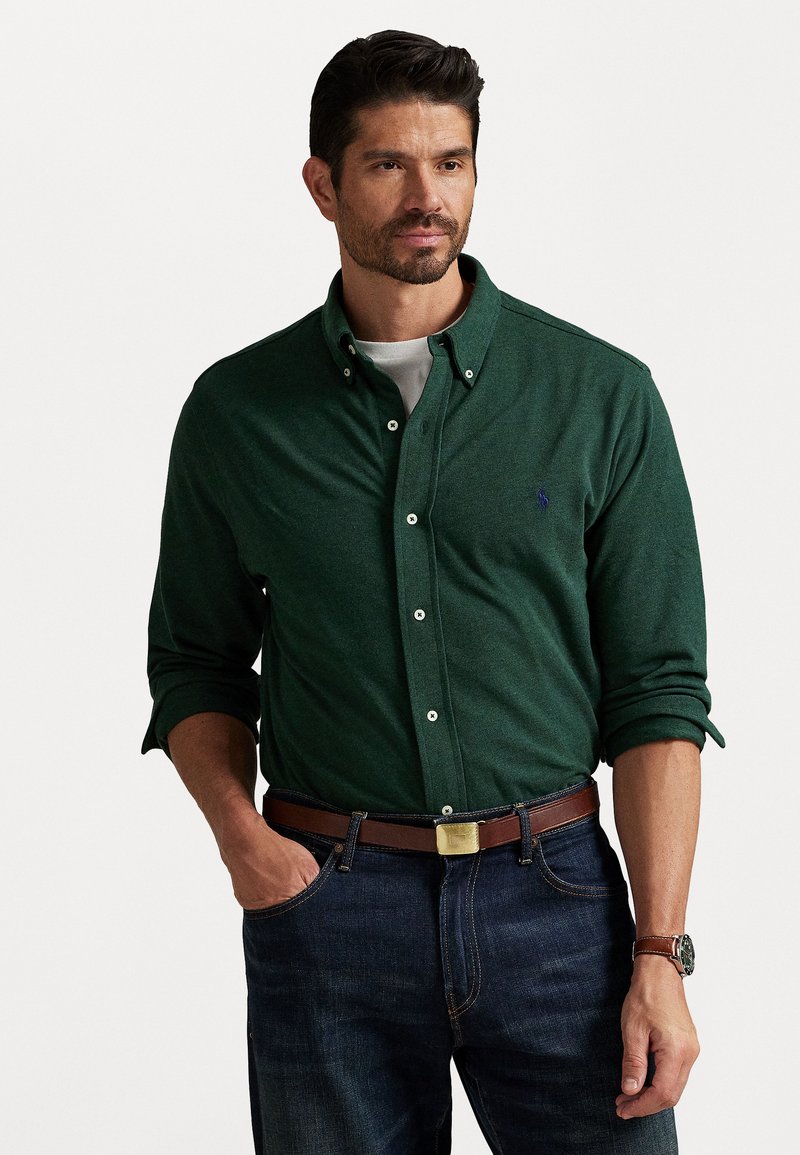 Mann med kort mørkt hår og skjegg iført mørkegrønn skjorte med knapper over hvit t-skjorte, blå jeans, brun belte og klokke, stående med hånden i lommen.