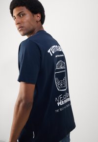 Marineblå bomull t-skjorte med en hvit grafisk illustrasjon av en drink og teksten "TUTTO BENE, NEGRONI PER FAVORE" på baksiden.