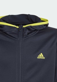 Felpa con cappuccio in poliestere blu navy con dettaglio giallo sul cappuccio e sul logo. Presenta un colletto alto e una texture liscia.