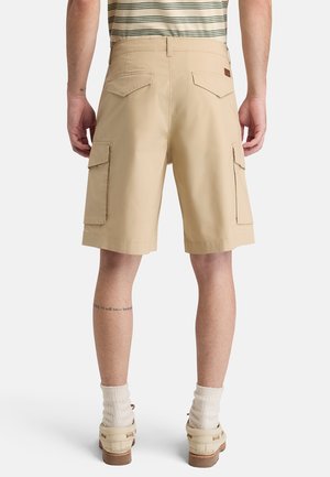 Mężczyzna ubrany w beżowe krótkie spodnie typu cargo, białe skarpetki i beżowe buty, widziany od tyłu na jednolitym tle.