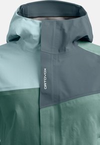 Waterproof jacket in shades of light blue and green. Features a high collar, a geometric design, and "ORTOVOX" logo in white. Smooth texture.