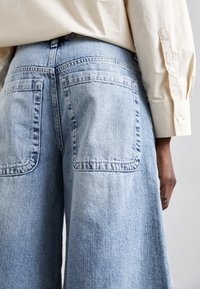 Pantalones de mezclilla de pierna ancha de color azul claro con grandes bolsillos traseros, que presentan un tejido texturizado y un diseño de cintura alta, parcialmente cubiertos por una camisa en color blanco roto.