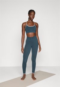 Giovane donna in piedi a piedi nudi su un tappeto da yoga, indossando un crop top atletico color teal scuro e leggings, davanti a uno sfondo chiaro e uniforme.