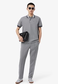 Completo grigio di polo e pantaloni con motivo a diamante, dettagli del colletto blu navy e logo verde. Il modello indossa occhiali da sole e porta una borsa nera.