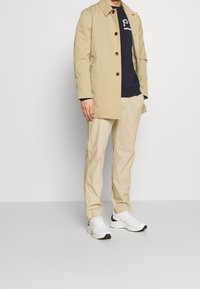 Trench beige claro con botones marrón oscuro, llevado sobre un sudadera azul marino, combinado con unos pantalones beige claro y zapatillas blancas.