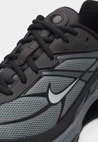 Κλείσιμο εικόνας ενός γκρι και μαύρου αθλητικού παπουτσιού με μαύρα κορδόνια και λευκό λογότυπο Nike Swoosh σε υφή από διχτυωτό υλικό.