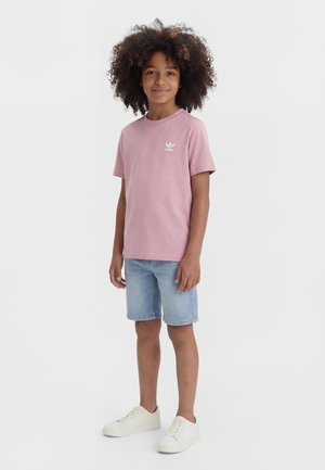 Barn med krøllet hår iført en lyserød Adidas t-shirt, lyse denimshorts og hvide sneakers, stående mod en hvid baggrund.