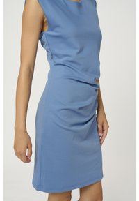 Robe bleue sans manches jusqu'aux genoux portée par une personne montrée des épaules jusqu'aux genoux sur un fond clair uni.