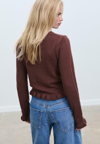 Maglione marrone lavorato a maglia con una texture a costine, caratterizzato da un orlo arruffato, abbinato a jeans azzurri larghi con tasche posteriori.