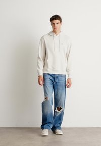 Helle beigen Kapuzenpullover mit Kängurutasche, kombiniert mit hellblauen, ausgewaschenen Weitbein-Jeans. Model trägt weiße Sneakers.