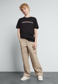 T-shirt nera con il testo "JORDAN" in bianco e rosso, abbinata a pantaloni beige dal fit ampio, indossata con sneakers chiare.