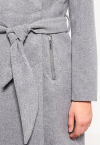 Abrigo de lana suave y gris con cinturón de lazo; cuenta con un bolsillo con cremallera en el lado izquierdo y un acabado texturizado.