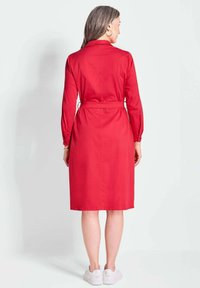 Rode jurk tot de knie met een getailleerde taille, lange mouwen en een kraag. Gemaakt van een soepele stof, met een strik aan de achterkant voor aanpassing.