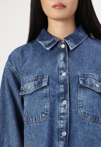 Camicia di denim di blu medio, con colletto, due tasche sul petto e bottoni argentati. Il tessuto ha un aspetto testurizzato e leggermente sbiadito.