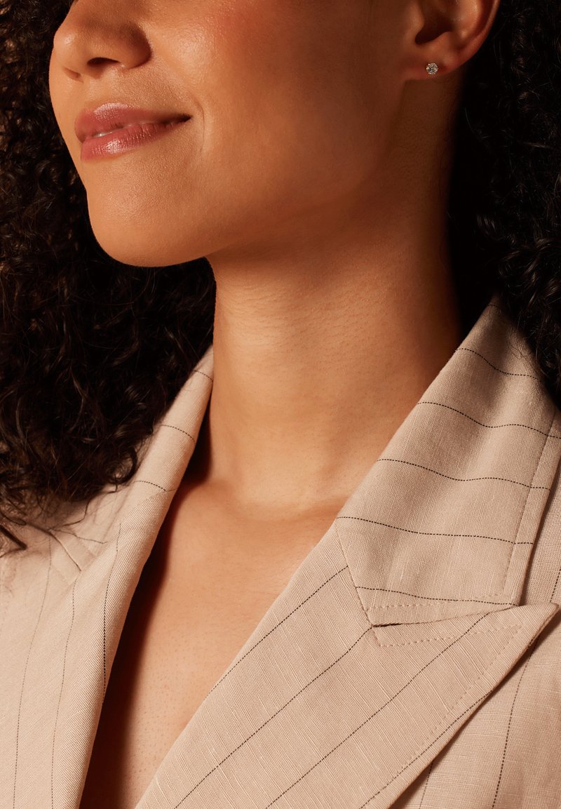 Gouden studs in de oren van een persoon met krullend haar, die een beige blazer met streepjes draagt. De stof heeft een gestructureerde afwerking.