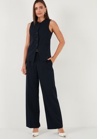 Vest bleumarin, croită impecabil, cu șase butoane, asortată cu pantalonii largi. Material neted. Pantofii albi cu toc adaugă un element de contrast.