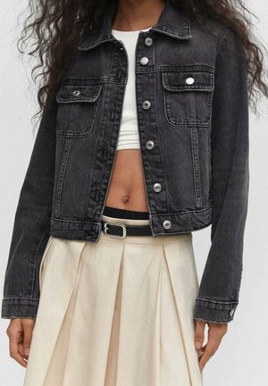 Kvinde iført en cropped sort denimjakke over en hvid crop top, kombineret med et beige plisseret skørt og sort bælte.