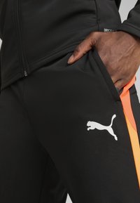Črne atletske hlače iz tkanine, ki odvaja vlago, z oranžnimi stranskimi detajli in belim logotipom Puma. Imajo stranske žep.