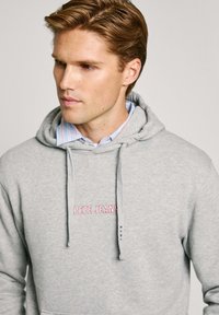 Sudadera gris con capucha ajustable, que presenta un logotipo rosa de "PEPE JEANS". Llevada sobre una camisa de cuello a rayas en azul claro y blanco.