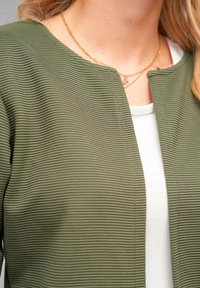 Olive green ribbed cardigan with a round neckline, featuring a textured surface and open front design. Worn over a white top.
