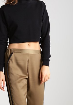 Kobieta ubrana w czarny krótki top z długim rękawem oraz beżowe spodnie z wysokim stanem i czarnymi paskami po bokach, stojąca na jednolitym tle.