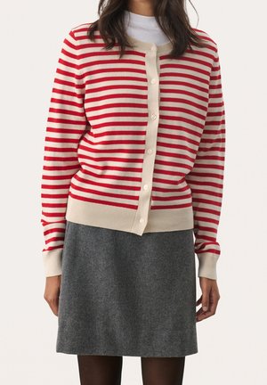 Femme portant un cardigan boutonné à rayures horizontales rouges et blanches et une jupe grise longueur genou, debout devant un fond uni.