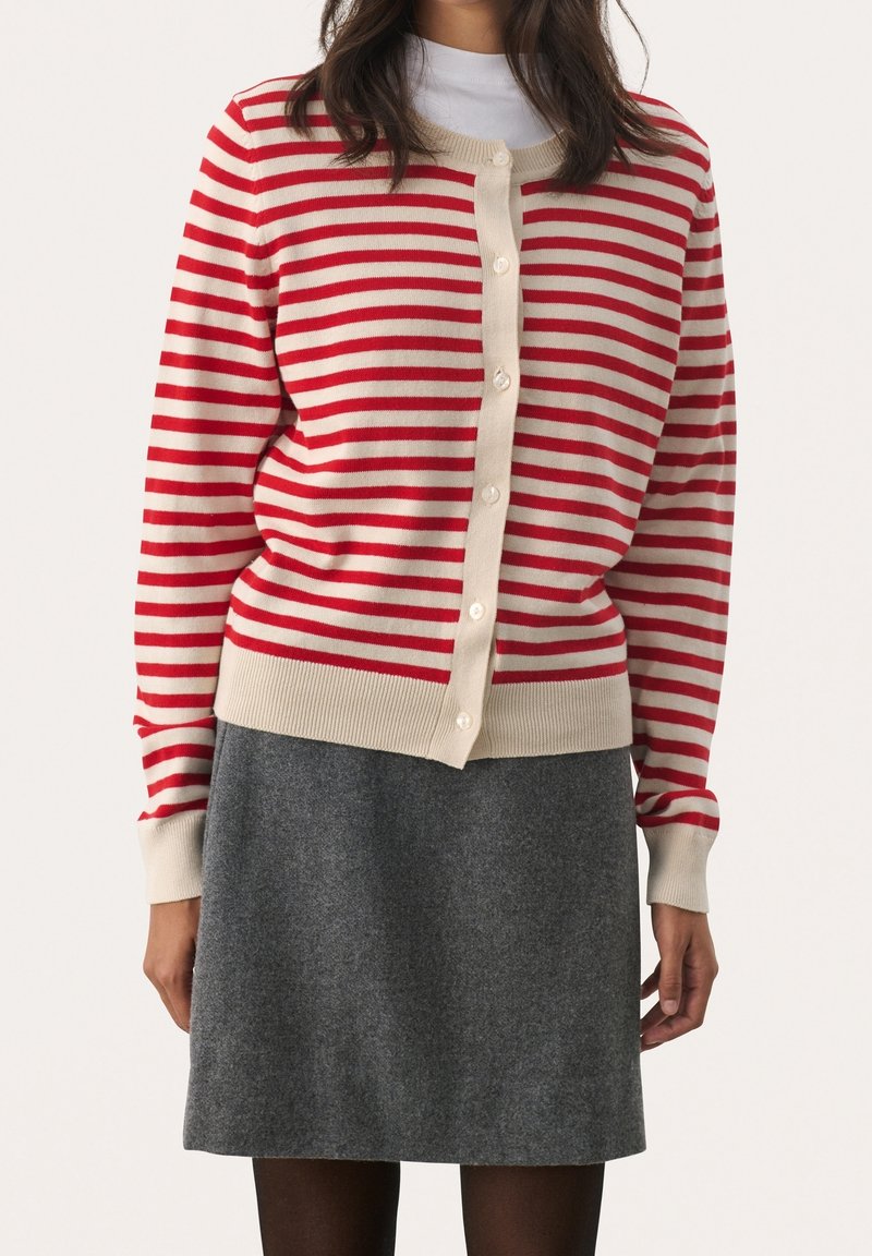 Femme portant un cardigan boutonné à rayures horizontales rouges et blanches et une jupe grise longueur genou, debout devant un fond uni.