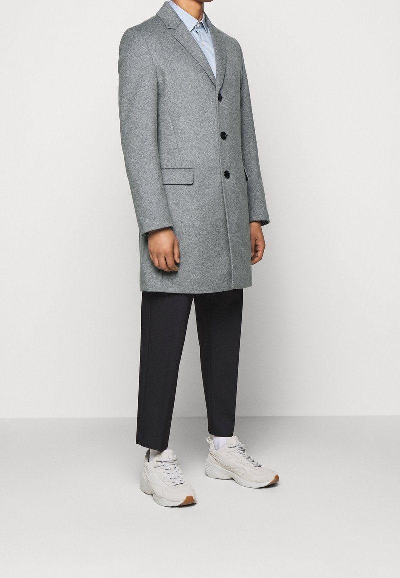 Un uomo che indossa un cappotto grigio lungo fino al ginocchio, pantaloni neri a vita alta, calze bianche e sneaker bianche chunky, in piedi su uno sfondo bianco uniforme.