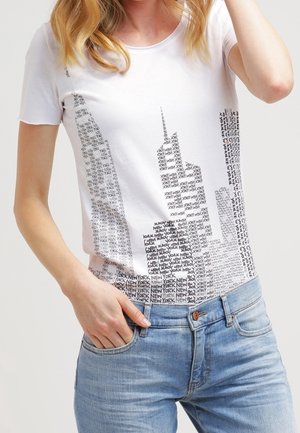 T-shirt blanc avec texte noir formant la silhouette de New York, porté avec un jean bleu clair par une personne aux cheveux blonds.