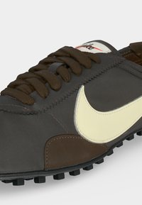 Σκούρο καφέ αθλητικό παπούτσι Nike με κρεμ χρώματος σήμα, χοντρές κορδόνια και υφή στην σόλα, εμφανιζόμενο από κοντινή πλευρική γωνία σε λευκό φόντο.