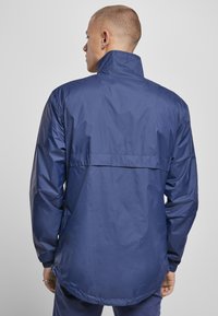 Mørkeblå vandtæt jakke med høj krave, lange ærmer, elastiske manchetter og en afslappet pasform. Glat, skinnende tekstur med en rygventilation.