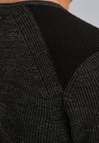 Close-up van zwart gebreid stof met variërende ribbelpatronen en gestructureerde steken die diagonale en verticale lijnen vormen.