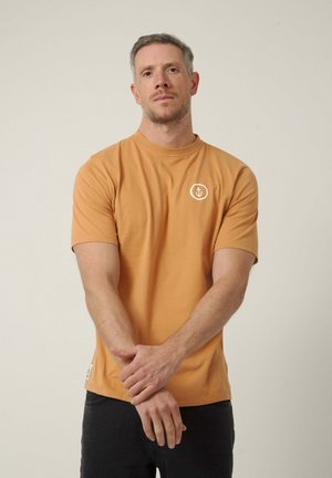 Hombre con cabello corto y barba incipiente, vistiendo una camiseta color mostaza con un emblema blanco de ancla, de pie contra un fondo claro y liso.