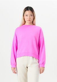 Young woman with long hair wearing a bright pink oversized sweatshirt and light beige pants, standing against a plain white background.