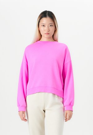 Jeune femme avec de longs cheveux portant un sweatshirt surdimensionné rose vif et un pantalon beige clair, se tenant devant un fond blanc uni.