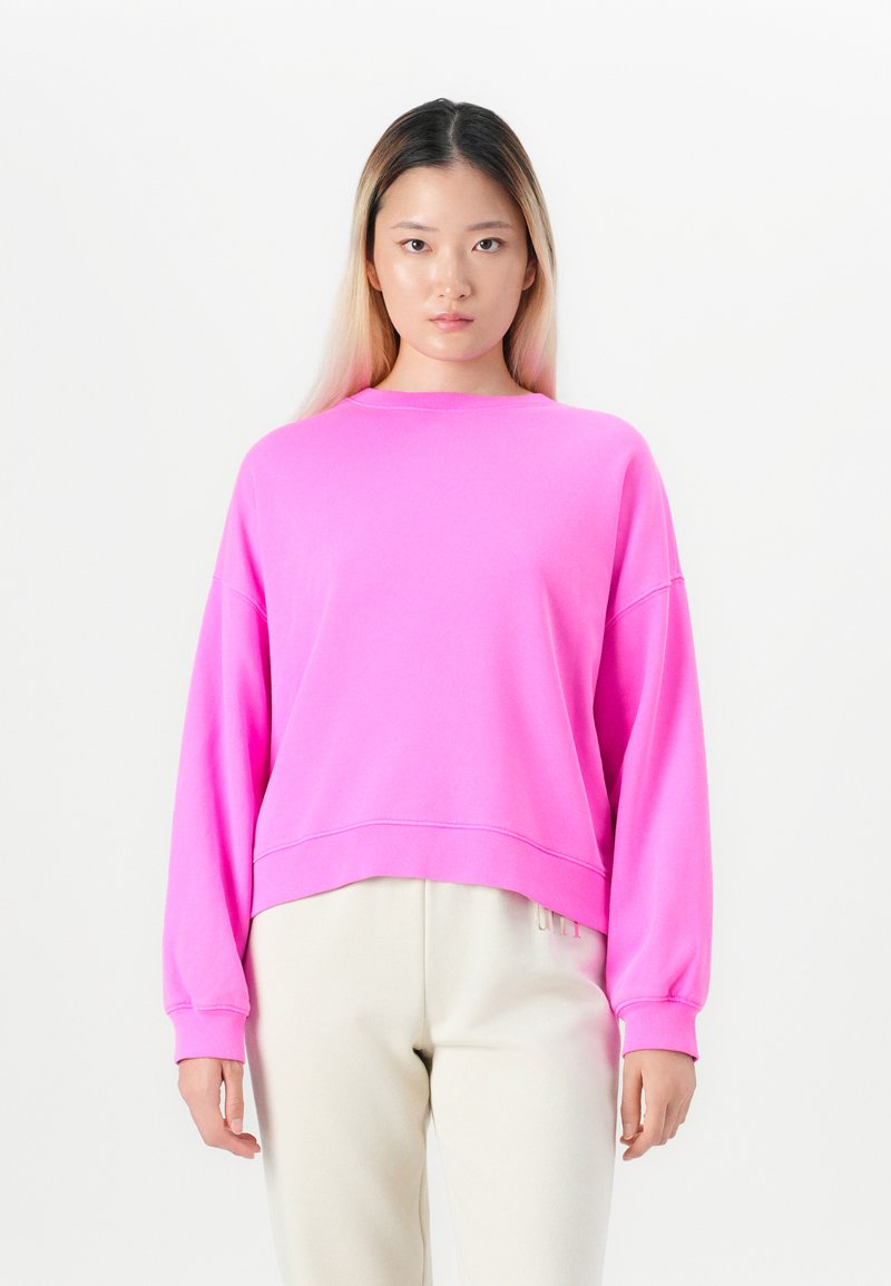 Young woman with long hair wearing a bright pink oversized sweatshirt and light beige pants, standing against a plain white background.