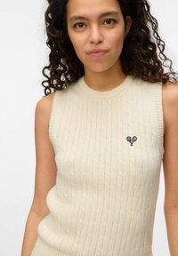 Top in maglia a coste di colore beige chiaro, con scollatura rotonda e un design ricamato di una racchetta da tennis sul lato sinistro del petto.