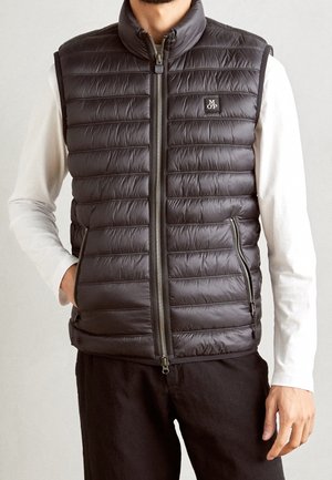 Uomo che indossa un gilet trapuntato nero con zip sopra una camicia bianca a maniche lunghe e pantaloni neri, con le mani nelle tasche del gilet.