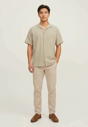Homme debout portant une chemise beige à manches courtes boutonnée, un pantalon beige clair et des chaussures marron sur un fond clair uni.