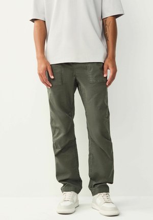 Person in olivgrünen Cargo-Shorts, weißen Sneakers und einem lockeren hellgrauen T-Shirt, das vor einem schlichten Hintergrund steht.
