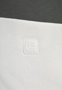 Vitt bomullstyg med en texturerad yta, med en grå överdel och en sydd logotypapplikation i fyrkantig form på den vita delen.