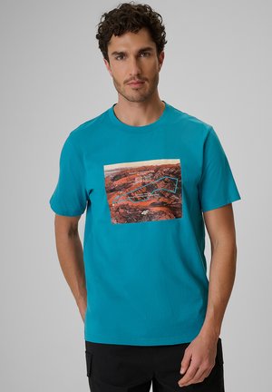 Mężczyzna z kręconymi włosami ubrany w turkusowy t-shirt z grafiką przedstawiającą pustynny krajobraz, tekstem i geometrycznym obrysem na froncie.